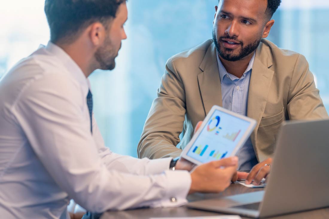 Two businessmen having a discussion in an office, with one showing financial graphs on a tablet.