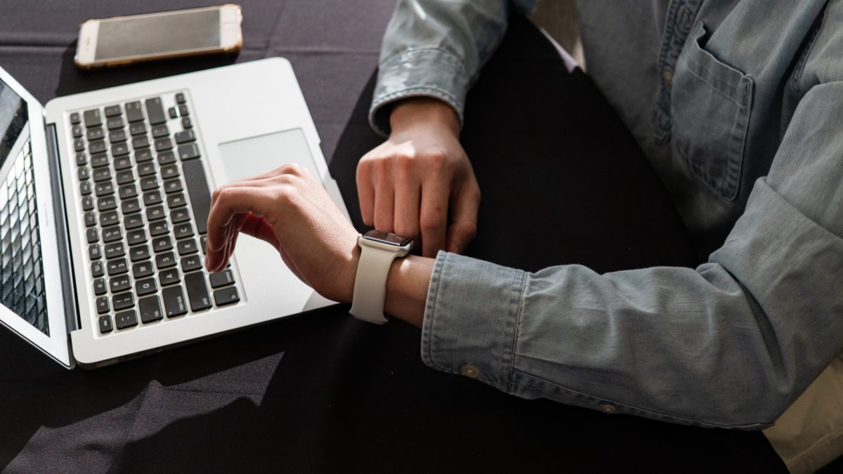 Seen from above, a man looking at his watch. His laptop and phone are on the table he's sitting at.