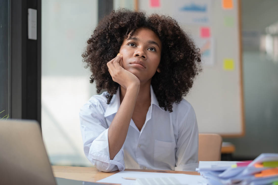 Woman in white shirt sitting at a desk, looking thoughtful.