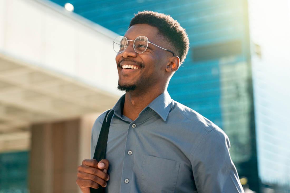 Smiling young man wearing glasses and a button-up shirt, walking outdoors with a backpack in a modern urban setting.